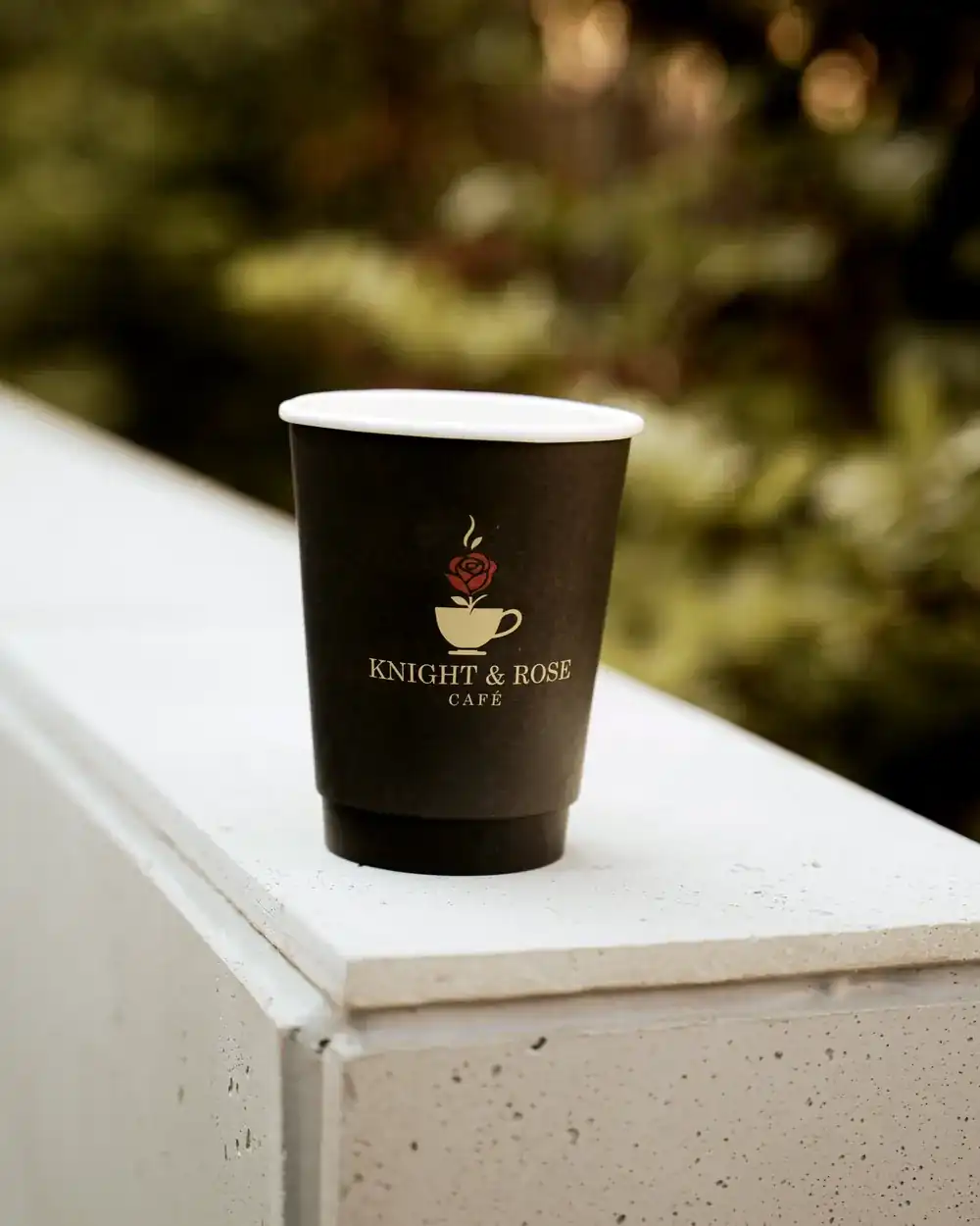 A Knight & Rose Café takeaway cup with its gold rose emblem resting on a stone ledge in afternoon light.